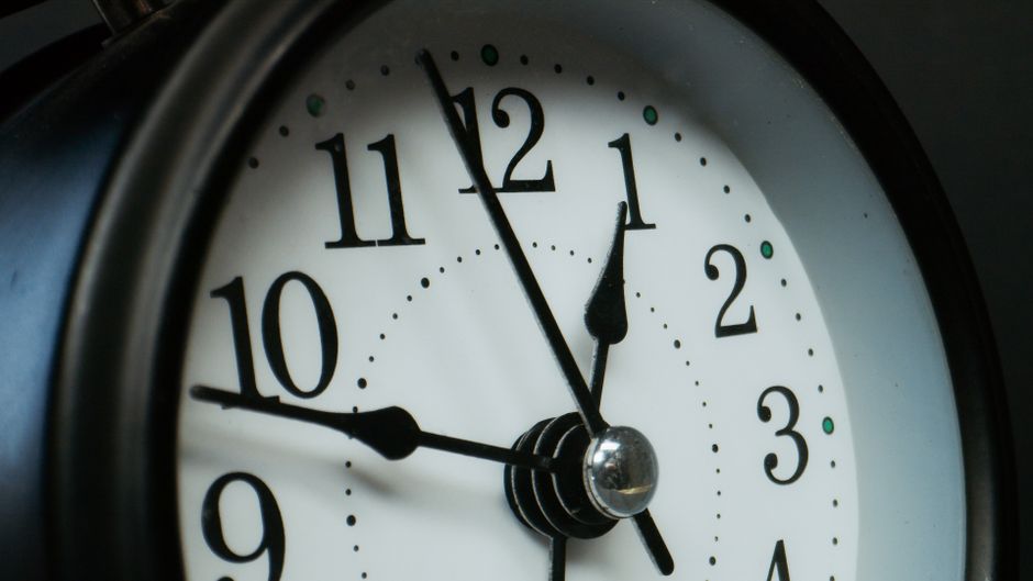 A close-up image of a round, analog wall clock featuring a white face with black Arabic numerals and black minute and hour hands, along with a thin black second hand. The clock shows the time as appro