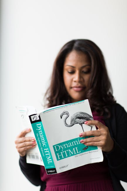 The woman, with long dark hair and wearing a black blazer over a burgundy top, is holding an open book titled 'Dynamic HTML' with a picture of a flamingo on the cover; she appears to be studying or re