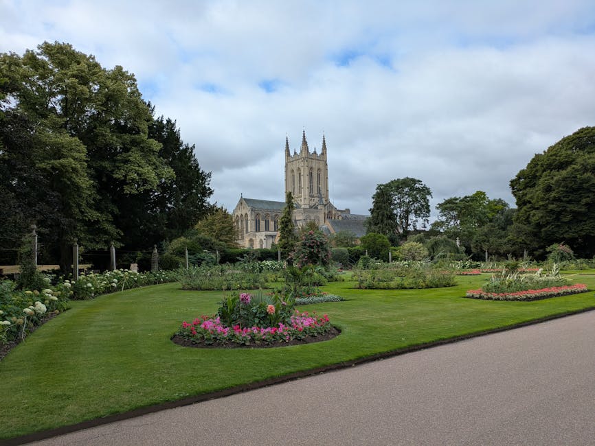 A scenic view of a well-maintained garden in front of a historic church, featuring lush green lawns with neatly trimmed grass, bordered by flower beds filled with colorful blooms such as pink and whit